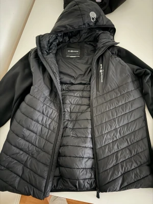 Svart pufferjacka från Sail Racing - Snygg svart pufferjacka från Sail Racing med huva och dragkedja. Jackan har quiltad design på framsidan och ärmar i slätare material. Perfekt för kalla dagar och har en sportig look med logga på bröstet.