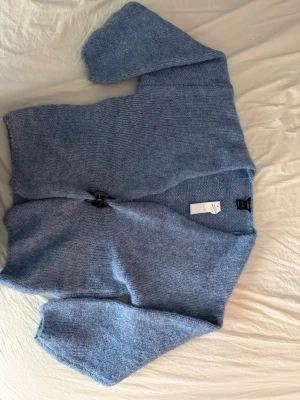 Blå stickad kofta från Lindex - Mysig blå stickad kofta från Lindex med v-ringning. Koftan har en avslappnad passform och långa ärmar. Perfekt att slänga över en t-shirt för en mysig och fin outfit. Helt oanvänd.