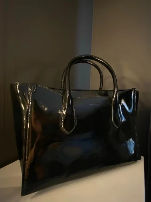 Svart lackad handväska från POLO Ralph lauren  - Säljer en svart, glansig handväska från POLO i lackat material. Väskan har två rejäla handtag och kommer även med ett axelremsband. Diskret logga framtill och minimalistisk design som ger en lyxig känsla.
