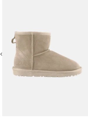  (FAKE UGGS) Beige fårskinnstofflor från Exani - Mysiga beige tofflor från Exani i klassisk modell med mjukt fårskinn på insidan och mockaliknande material på utsidan. Tofflorna har en låg skaft och en rejäl sula som ger bra grepp. Perfekta för att hålla fötterna varma under kalla dagar.