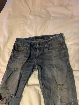 Blå slitna jeans från Replay - Säljer ett par blå jeans från Replay med slitningar och hål på benen. Klassisk femficksmodell med knappgylf och smal passform. Jeansen har en cool, tvättad look och är tillverkade i denim.