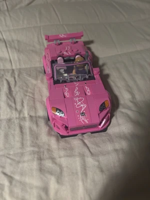 Rosa LEGO sportbil med dekaler - Säljer en rosa LEGO sportbil med vita dekorativa mönster på motorhuven och spoilern. Bilen har öppet tak, detaljerade strålkastare och två minifigurer i förarsätet. Perfekt för dig som gillar byggsatser och coola bilar.