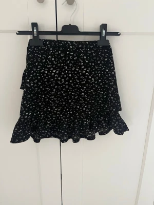 Svart blommig volangkjol - Supersöt svart kortkjol från Kappahl med små vita blommor och volangdetaljer. Perfekt att styla med en enkel topp eller hoodie.Säljer pga för liten.Storlek 146-152/XS