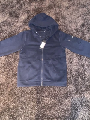 Marinblå zip-hoodie från Sail Racing - Snygg marinblå hoodie från Sail Racing med dragkedja framtill, två fickor och en extra ficka med dragkedja på ärmen. Klassisk huva och diskret Sail Racing-logga på ärmen. Perfekt för en avslappnad och sportig stil och grisch pris kan förhandlas. 