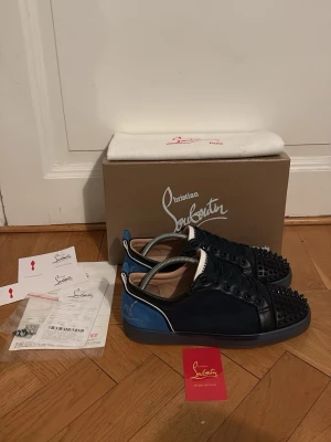Christian Louboutin mörkblå sneakers - Säljer ett par mörkblåa sneakers från Christian Louboutin med blå detaljer och svarta nitar på tån. Skorna har en cool kontrast mellan mocka och läder, samt vita och blå inslag på hälen. Perfekta för dig som vill sticka ut med exklusiv streetstyle. Det är en sällsynt colorway. Det är storlek 43. Låda, dustbag, kvitton, extra spikar, Louboutin kort medföljer (allt på bilderna). 