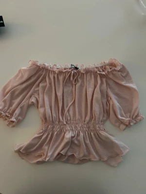 Ljusrosa offshoulder blus med volang - Supersöt ljusrosa offshoulder blus med volangkant och smock i midjan. Toppen har korta puffärmar, knyt framtill och en luftig, flowy passform. Perfekt för dig som gillar romantisk och feminin stil. Aldrig använd! 