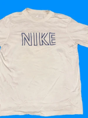 Vit Nike t-shirt med blå logga - Vit t-shirt från Nike med blå, grafisk logga på framsidan och klassisk Nike swoosh på baksidan. T-shirten har rund hals och korta ärmar, perfekt för dig som gillar sportiga och stilrena plagg.
