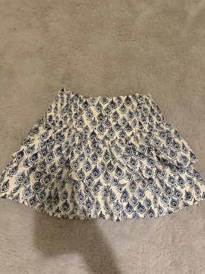 Vit och blå mönstrad kjol från Zara - Kort kjol från Zara i vitt med blått bladmönster. Kjolen har lite stretch i midjan aldrig  använt strolek 152 