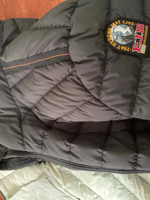 Svart dunjacka från Parajumpers - Säljer en svart dunjacka från Parajumpers med snyggt mönstrad quiltning och patch på bröstet. Jackan har dragkedja med orange detalj och är perfekt för kalla vinterdagar. Materialet är slitstarkt och håller värmen riktigt bra.