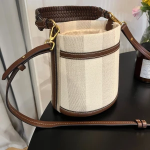 Zara väska  - Snygg bucket bag i beige bomullstyg med breda ränder och bruna skinn-detaljer. Väskan har justerbar axelrem, flätat handtag och stängs med snörning upptill. Perfekt för dig som vill ha en trendig och rymlig väska med stilrena detaljer Slutsåld på Zara