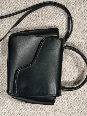 Svart handväska från ATP Atelier - Snygg svart handväska från ATP Atelier i slätt läder. Väskan har en unik asymmetrisk lockdesign, magnetlås och både axelrem och handtag. Finns små tecken på användning men annars är den i superfint skick!💞💞