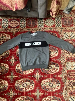 Grå sweatshirt från Nike - Snygg grå sweatshirt från Nike med svart band och vit logga över bröstet. Tröjan har rund halsringning och långa ärmar. Tillverkad i mjukt material som passar perfekt till en avslappnad stil.