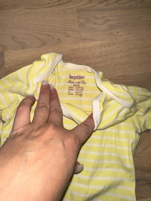 Gulrandig långärmad body från Lupilu - Söt långärmad body från Lupilu i storlek 86/92. Den är gul med vita ränder och har vita kantband. Tillverkad i mjuk bomull, perfekt för små barn. Praktiska tryckknappar i grenen gör den enkel att ta av och på.