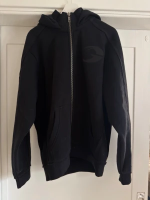 Svart zip hoodie från gasp - Svart hoodie med dragkedja från gasp. Stor logga i svart ton på ton på bröstet, två fickor framtill och ribbade muddar vid ärmslut och nederkant. Tillverkad i mjukt sweatshirtmaterial och har en klassisk huva. Tröjan är helt ny och inte använd