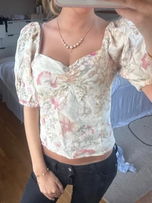 Blommig puffärm blus i ljusa toner - Söt blus med puffärmar och v-ringning i ljusa färger med blommigt mönster. Toppen har en croppad passform och är tillverkad i linne, perfekt för dig som gillar romantiska och trendiga plagg.