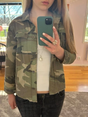 Camouflage jeansjacka från Zara - Snygg jeansjacka från Zara i klassiskt camouflagemönster. Jackan har krage, knäppning framtill och två bröstfickor samt två sidofickor.  Den har en rå kant nertill för en cool look.