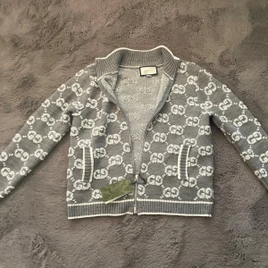 Gucci cardigan  - Snygg grå kofta från Gucci med hel dragkedja och ribbade muddar. Koftan har ett vitt GG-logomönster över hela plagget och två fickor framtill. Tillverkad i mjukt stickat material och har en klassisk krage. Perfekt för dig som gillar exklusiva märken. Och perfekt till vara stilig till sommaren! Vid snabb affär kan pris diskuteras!