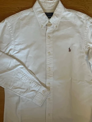Vit oxford skjorta från Polo Ralph Lauren - Klassisk vit oxford skjorta från Polo Ralph Lauren med broderad logga på bröstet. Skjortan har lång ärm, knappar framtill och en stilren krage. Perfekt för en clean och tidlös look.