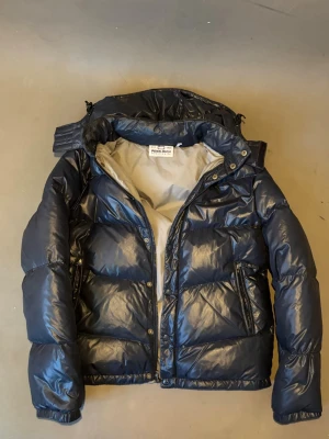 Woolrich pufferjacka - Snygg mörkblå pufferjacka från woolrich med glansig finish och avtagbar huva. Jackan har dragkedja och tryckknappar framtill, två snedställda fickor med dragkedja och ljusgrått innerfoder. Perfekt för kalla vinterdagar.