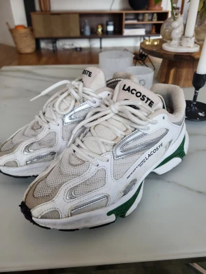 Vita sneakers från Lacoste - Snygga vita sneakers från Lacoste med mesh och läderdetaljer, silverfärgade inslag och gröna partier på sulan. Klassisk logga på plösen och sidan. Skorna har en sportig vibe och är perfekta för dig som gillar streetstyle.