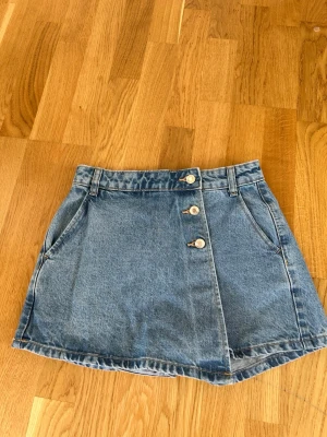 Shortskjol från Zara - Snygg blå denim shortskjol från Zara med omlott-look framtill och tre dekorativa knappar. Klassisk femficksdesign med två framfickor och två bakfickor. Skortsen har bälteshällor och är gjord i ett robust jeanstyg. Perfekt för en avslappnad och trendig stil. Kostar 350kr nypris på Zara. Säljer för 250kr då de är försmå. Använda 3 gånger. 