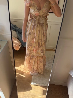 Blommig långklänning med volang - Romantisk långklänning i ljusa toner med blommigt mönster. Klänningen har volangdetaljer, v-ringning, öppen rygg och knyt i midjan. Ärmlös med tunna axelband och tillverkad i ett lätt, luftigt material som passar perfekt för sommarens varma dagar.