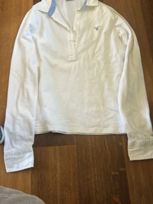 Vit långärmad henley från Gant - Klassisk vit henley-skjorta från Gant med lång ärm och ljusblå detalj på insidan av kragen. Skjortan har knappar framtill och en liten broderad logga på bröstet. Perfekt för en clean och stilren look.