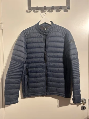 Mörkblå dunjacka från Replay - Snygg mörkblå pufferjacka från Replay med quiltad design och hög krage. Jackan har dragkedja framtill, två sidofickor med dragkedja och Replay-logga på kragen. Perfekt för kyliga dagar och enkel att matcha till olika outfits. Enda defekten är att Y ett som är på dragkedjan är borta, alltså klisterlappen som sitter ovanpå men man ser fortfarande att det står REPLAY. Andra är att man kan se lite slitage på knappar men då ska man kolla noga. MVH Liam/Brakansgarderob