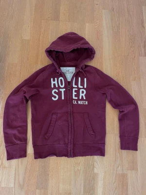 Hollister zip hoodie - Vinröd zip hoodie från Hollister | Storlek S | Två väldigt små hål på ena armen som inte märks av vid användning (se bilder), annars Fint skick✅| 