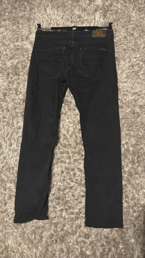 Nudie jeans - Säljer ett par svarta jeans från nudie jeansmed klassisk femficksdesign och raka ben. Jeansen har en läderpatch bak i midjan och är tillverkade i ett mjukt denim-material. Perfekta för dig som gillar en enkel och stilren look. LEAN DEAN
