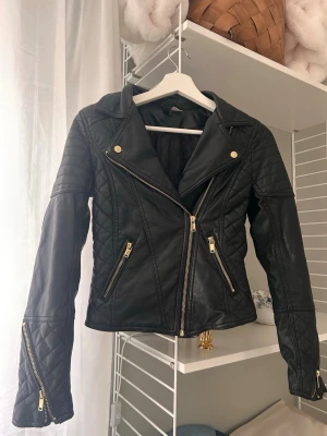 Svart biker skinnjacka med gulddetaljer - Snygg svart skinnjacka i bikerstil med guldfärgade dragkedjor och knappar. Jackan har quiltade detaljer på axlar och ärmar samt snedställd dragkedja framtill. Perfekt för en cool och trendig look.