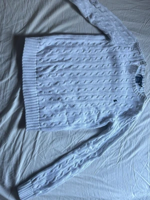 Vit kabelstickad tröja Ralph Lauren - Går att diskutera priset. Vit kabelstickad tröja från Ralph Lauren med rund hals och broderad mörkblå logga på bröstet. Tröjan har långa ärmar och ribbade muddar vid ärmslut och nederkant. Materialet är mjukt och skönt, perfekt för en stilren look. 