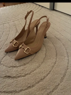 Beige slingback pumps med gulddetaljer - Säljer ett par stilrena beige pumps med öppen häl och justerbar rem. Skorna har spetsig tå och snygga guldfärgade metalldetaljer framtill. Klacken är medelhög och skon är tillverkad i slätt material som ger en elegant look.