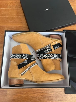 AMIRI cowboy boots med bandana - Snygga cowboy boots från AMIRI i ljusbrunt mocka med svartvit bandana-detalj och silverkedja runt skaftet. Boots med träklack och cool design som sticker ut. Perfekt för dig som vill ha något unikt och trendigt.