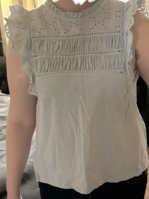 Ljusblå  ärmlös blus med spetsdetaljer - Söt ljusblå blus med broderade spetsdetaljer och volang vid axlarna. Hög hals med rynk och dekorativa hålmönster upptill. Blusen är ärmlös och har en luftig passform, perfekt till jeans eller kjol. Materialet känns mjukt och lätt.