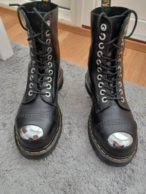Svarta Dr. Martens kängor med ståltå - Säljer ett par svarta Dr. Martens kängor i läder med ståltå och grov sula. Kängorna har högt skaft, snörning med metallhål och den klassiska gula sömmen runt sulan. Perfekta för dig som vill ha en tuff och robust stil.
