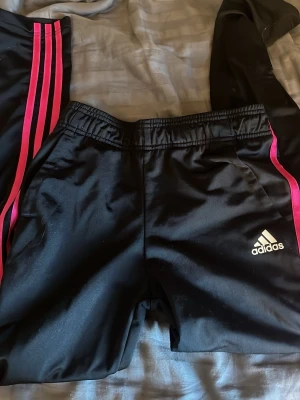 Svarta Adidas träningsbyxor med rosa ränder - Snygga svarta träningsbyxor från Adidas med klassiska rosa ränder längs sidorna och vit Adidas-logga på ena benet. Byxorna har resår i midjan och är tillverkade i ett glansigt, sportigt material som passar perfekt till träning eller chill.