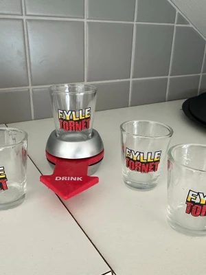 Fylletornet drickspel med glas - Fylletornet drickspel med fyra shotglas i glas med tryckt text. Spelet har en röd och silverfärgad snurrplatta med pil och texten 'DRINK'. Perfekt för förfesten eller spelkvällen med kompisarna.