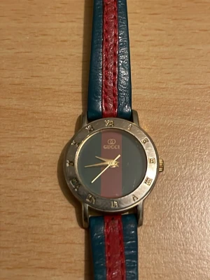 Gucci klocka med blått och rött läderband - Snygg klocka från Gucci med klassiskt blått och rött läderband. Urtavlan har en vertikal röd och blå rand och guldfärgade visare. Boetten är i metall med romerska siffror runt om. Perfekt accessoar för dig som gillar lyxig och unik stil.