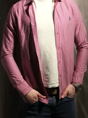 Rosa randig skjorta från Ralph Lauren - Säljer en snygg rosa skjorta med vita ränder från Polo Ralph Lauren. Skjortan har klassisk krage, knappar framtill och ett blått broderat logomärke på bröstet. Perfekt för dig som vill ha en stilren och trendig look.