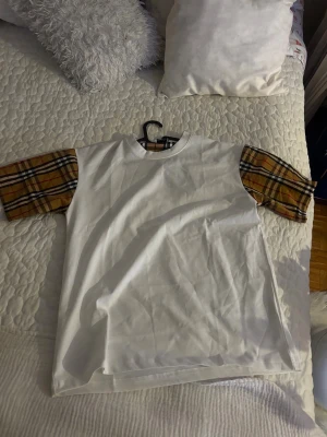 Burberry t shirt  - Vit t-shirt med korta ärmar i klassiskt brunt, svart och gult rutigt mönster. T-shirten har en rak passform och rund halsringning. Perfekt för dig som vill sticka ut med en unik detalj på en annars enkel t-shirt.