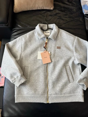 Grå zip-jacka från Miu iu - Snygg grå jacka från MiuMiu med dragkedja framtill och klassisk krage. Jackan har två fickor på sidorna och broderad MiuMiu-logga på bröstet. Perfekt för dig som gillar stilren och enkel design.