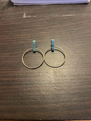 Guldiga örhängen med blå detalj - Säljer ett par stilrena örhängen i guldfärgad metall med stora runda ringar och en ljusblå detalj upptill, dem är från H&M. Endast testade men aldrig använda liksom. Skriv om du har frågor eller undrar något!