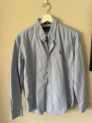 Ljusblå skjorta från Ralph Lauren - Klassisk ljusblå skjorta från Ralph Lauren med button-down krage och broderad logga på bröstet. Skjortan har långa ärmar och vita knappar framtill. Perfekt till jeans eller chinos för en clean och stilren look. Storlek L men är som S/M