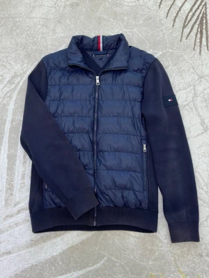 Marinblå Tommy Hilfiger jacka - Granatowa, lekka kurtka marki Tommy Hilfiger w rozmiarze M. Kurtka ma stylowy krój z pikowanym przodem ze 100% poliamidu i miękkimi, dzianinowymi rękawami zapewniającymi wygodę. Zapinana na zamek błyskawiczny z przodu i boczne kieszenie zapinane na zamek. Idealna jako kurtka przejściowa na wiosnę i jesień.  Uwaga: niektóre części kurtki mają przebarwienia (patrz zdjęcie), co ma wpływ na cenę. Poza tym w dobrym, używanym stanie.  Marinblå lättviktsjacka från Tommy Hilfiger i storlek M. 