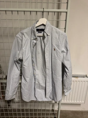 Randig skjorta från Ralph Lauren - Klassisk vit och blå randig skjorta från Ralph Lauren i slim fit-modell. Skjortan har långa ärmar, knappar framtill och den ikoniska broderade loggan på bröstet. Tillverkad i mjuk bomull som känns skön mot huden.