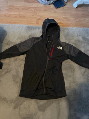 Svart hoodie från The North Face - Mörkgrå hoodie från The North Face med hel dragkedja, röda detaljer på dragkedjorna och logga på bröstet. Materialet är en mix av mjuk fleece och förstärkta partier på axlar och armar. Perfekt för dig som gillar en sportig och stilren look.
