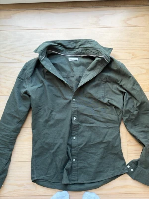 Mörkgrön skjorta med knappar - Snygg mörkgrön skjorta i bomull med klassisk krage och knappar hela vägen framtill. Skjortan har lång ärm och en clean, stilren look som passar till jeans eller chinos. Perfekt för dig som gillar enkel men trendig stil.