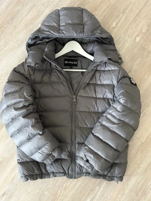 Grå pufferjacka från Bvaldi - Snygg grå pufferjacka från Bvaldi med stor huva och quiltad design. Nypris ligger på 2999kr. Jackan har dragkedja framtill, ribbade muddar och en patch på ärmen. Perfekt för kalla dagar och riktigt skön att bära. Materialet är glansigt och ger en modern look.