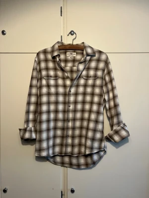 Rutig skjorta från American Eagle - Riktigt snygg rutig skjorta från American Eagle i beige, grå och vit. Skjortan har klassisk krage, knappar framtill och två bröstfickor med lock. Långärmad modell som är perfekt att bära öppen eller stängd. Materialet känns mjukt och bekvämt.